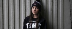 Teenage girl with beenie and sweatshirt that says "TIME." She is not smiling and leadning against a corrugated concreate wall.